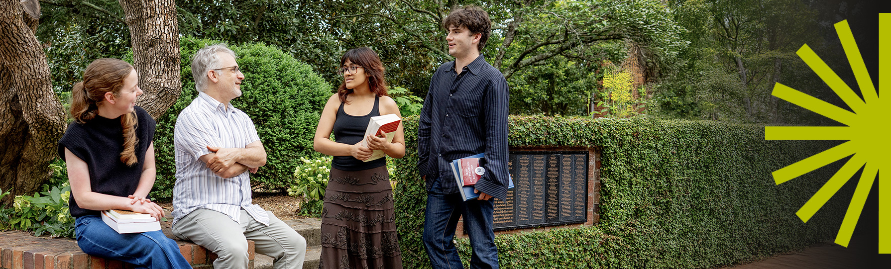 Two students standing and one sitting on a bench with their professor outside in a garden