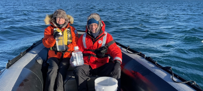 photo of two people in a small boat in water, snow-capped land in background