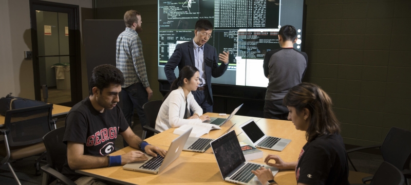 photo of six people in a room with laptop and projection screen, table