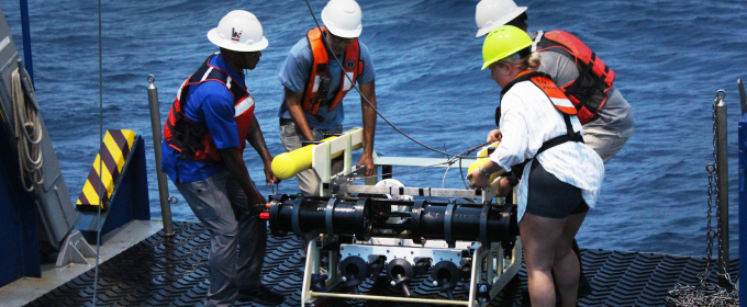 photo off four people with large device in deck of ship, day, water in background